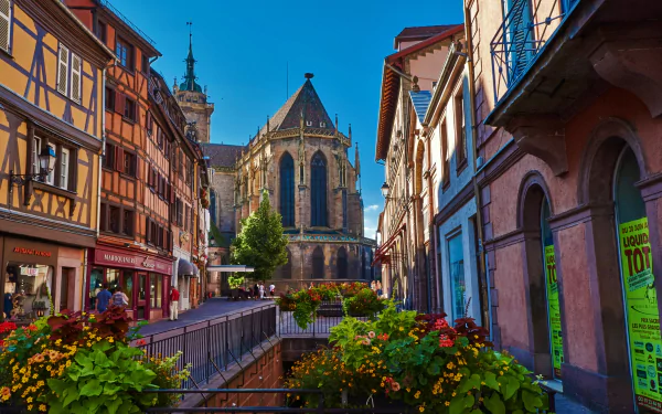 Colorful traditional buildings line a street in Colmar, France, with a historic church and vibrant flower boxes under a clear blue sky in this HD desktop wallpaper.