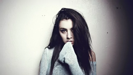 HD PC desktop wallpaper: moody portrait of a young female music artist in an oversized sweater against a pale wall, staring with a contemplative gaze.