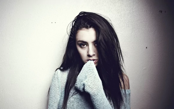 HD PC desktop wallpaper: moody portrait of a young female music artist in an oversized sweater against a pale wall, staring with a contemplative gaze.