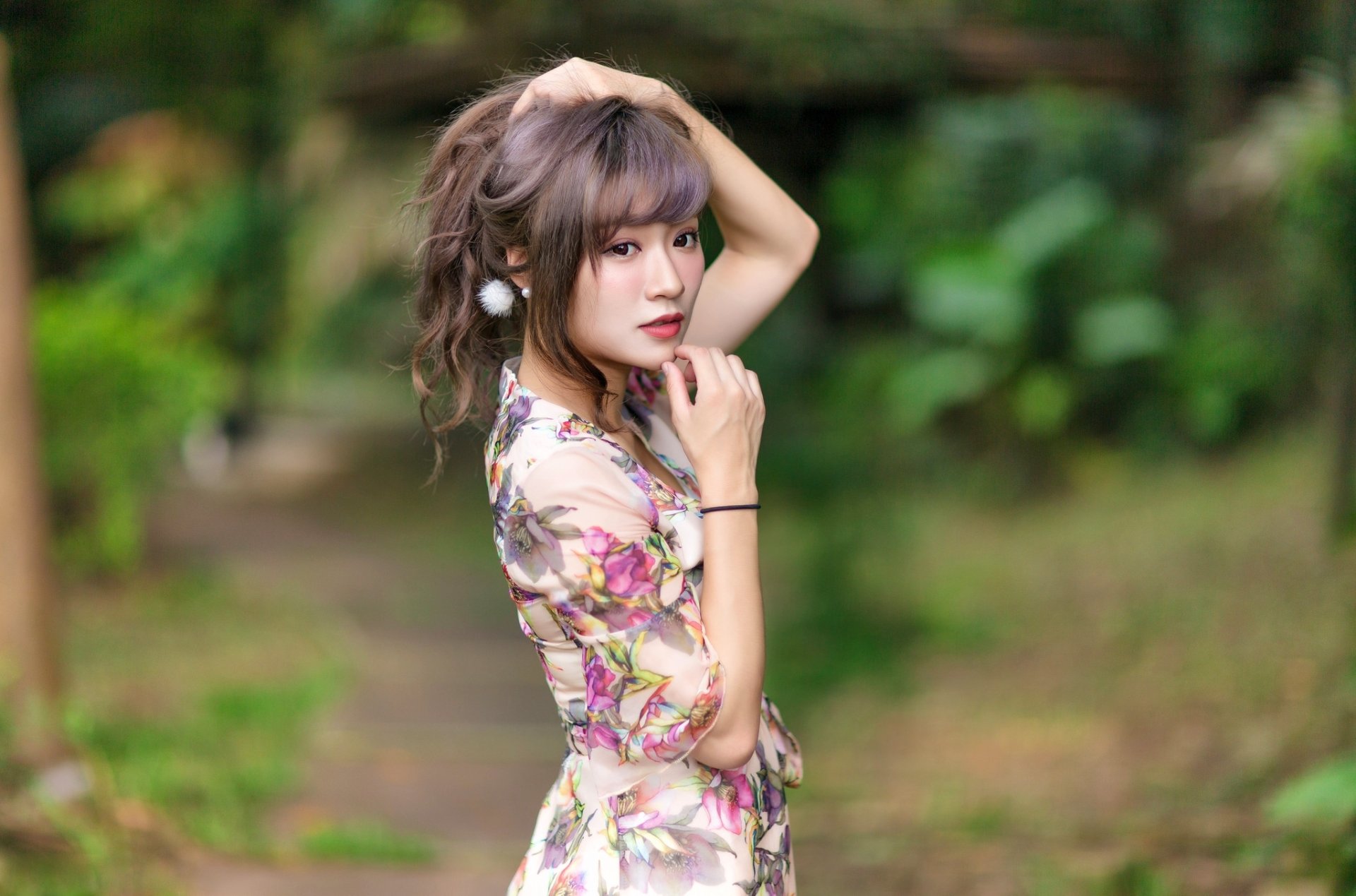 HD wallpaper featuring an Asian brunette woman with brown eyes in a floral dress. She poses gracefully outdoors with a blurred green background, highlighting the depth of field.