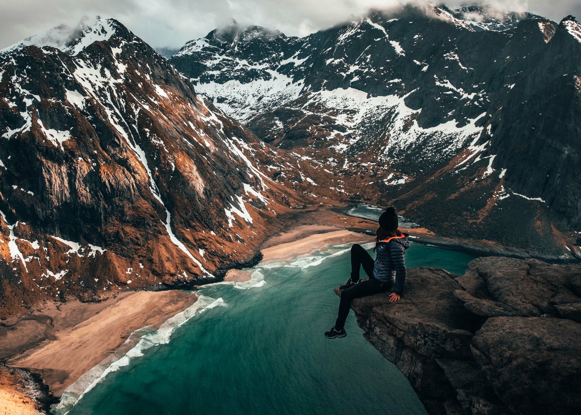 A woman sits on a rocky cliff overlooking a turquoise mountain lake surrounded by snow-dusted peaks, capturing a serene and contemplative mood in this HD desktop wallpaper.