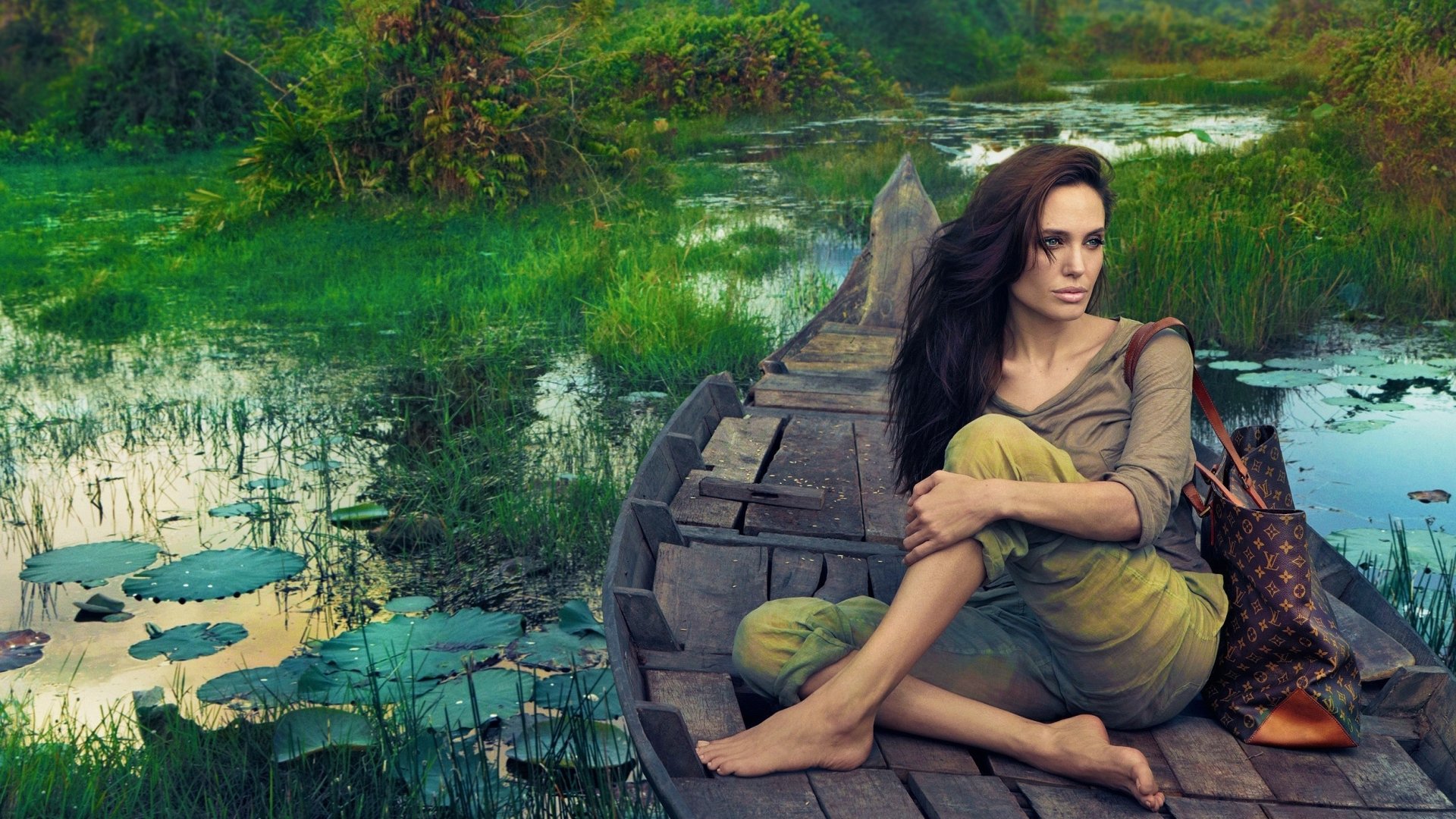 HD desktop wallpaper featuring American actress and celebrity Angelina Jolie sitting on a wooden boat amidst a tranquil, green, lotus-filled water scenery.