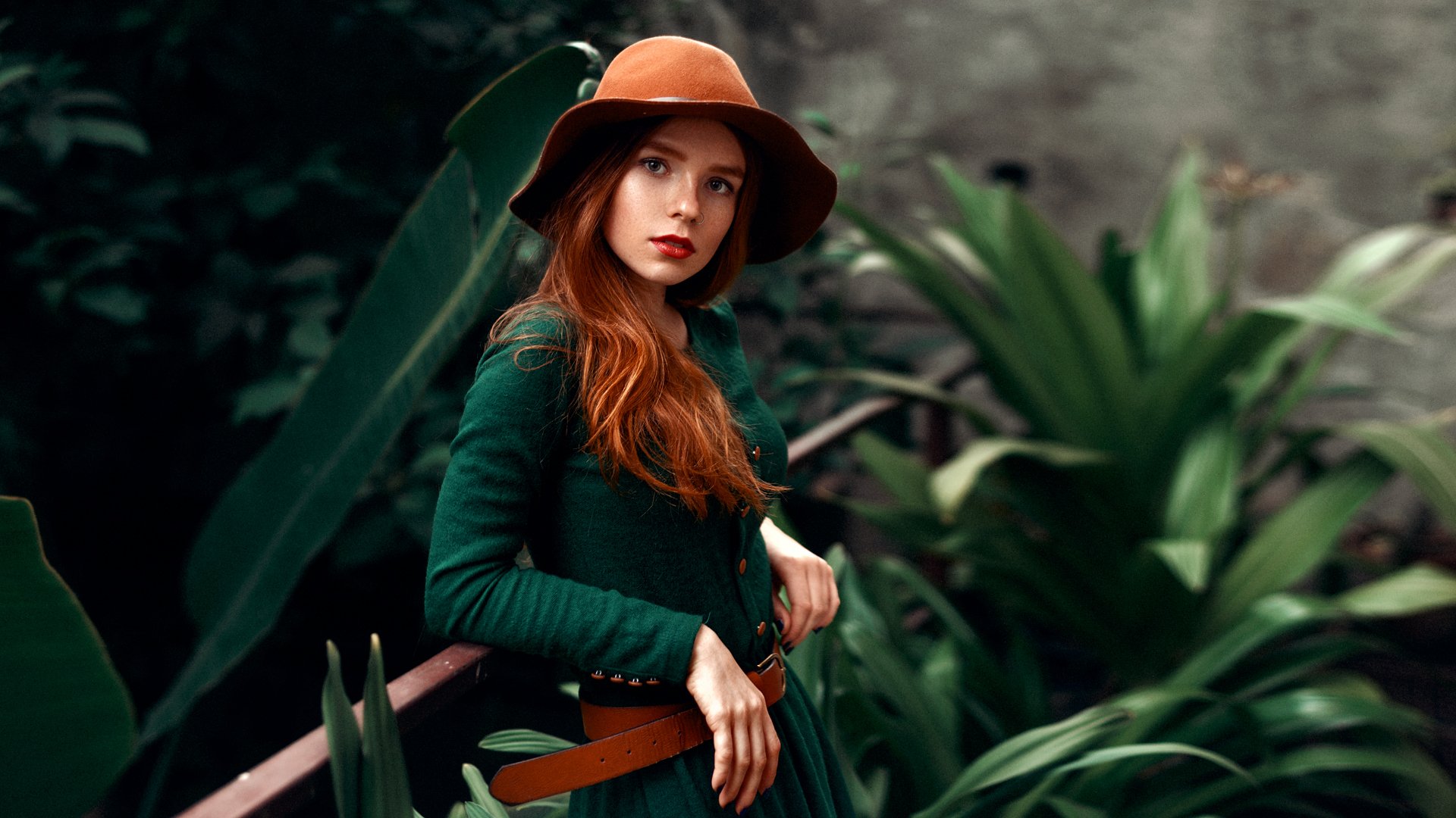 HD desktop wallpaper of a redhead woman wearing a brown hat, green dress with a belt, and lipstick, captured with depth of field against a lush, blurred foliage background.
