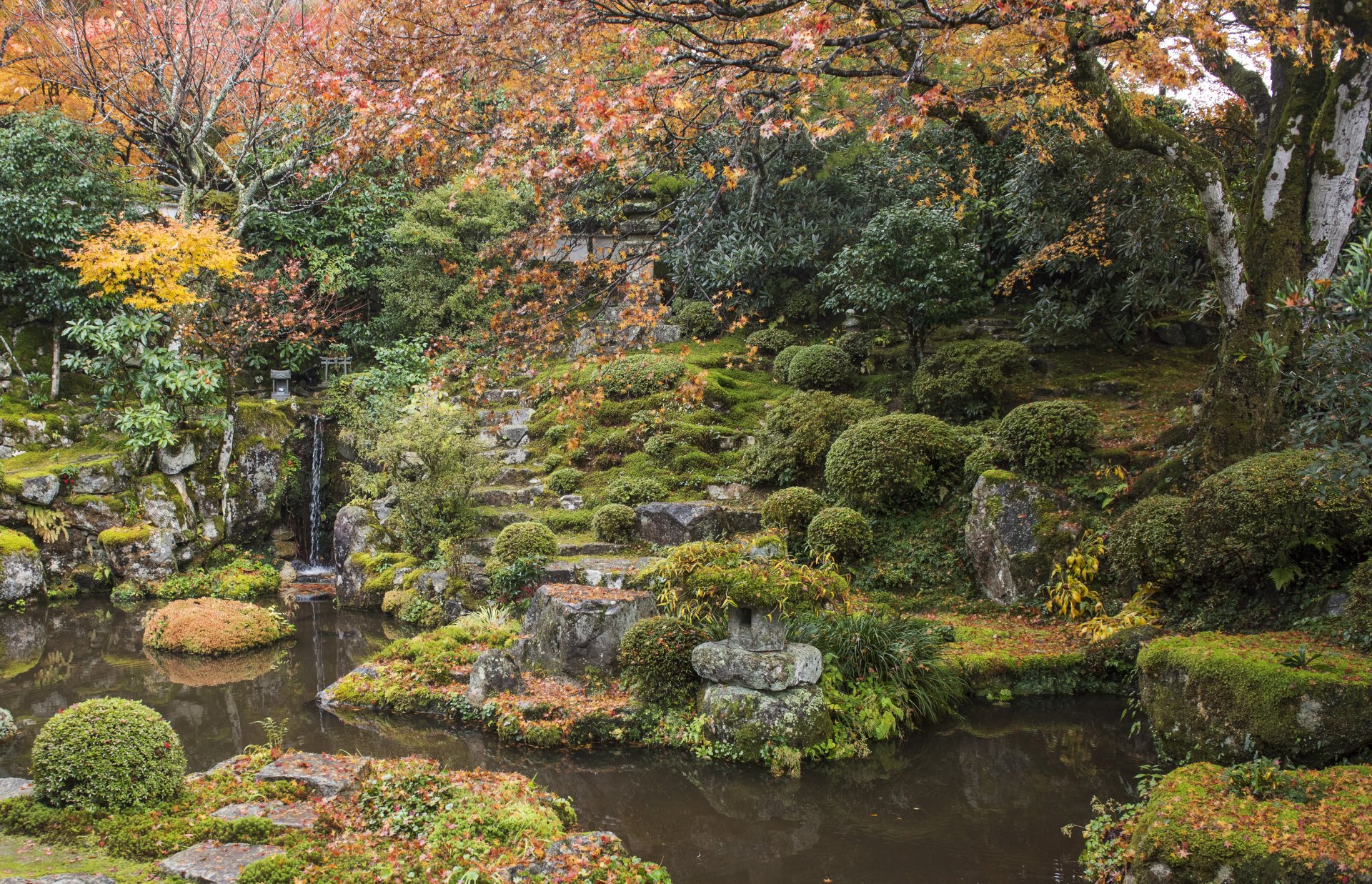 4K Ultra HD PC desktop wallpaper: tranquil man-made Japanese garden in fall — mossy stones, sculpted shrubs, small waterfall and reflective pond framed by autumn foliage.