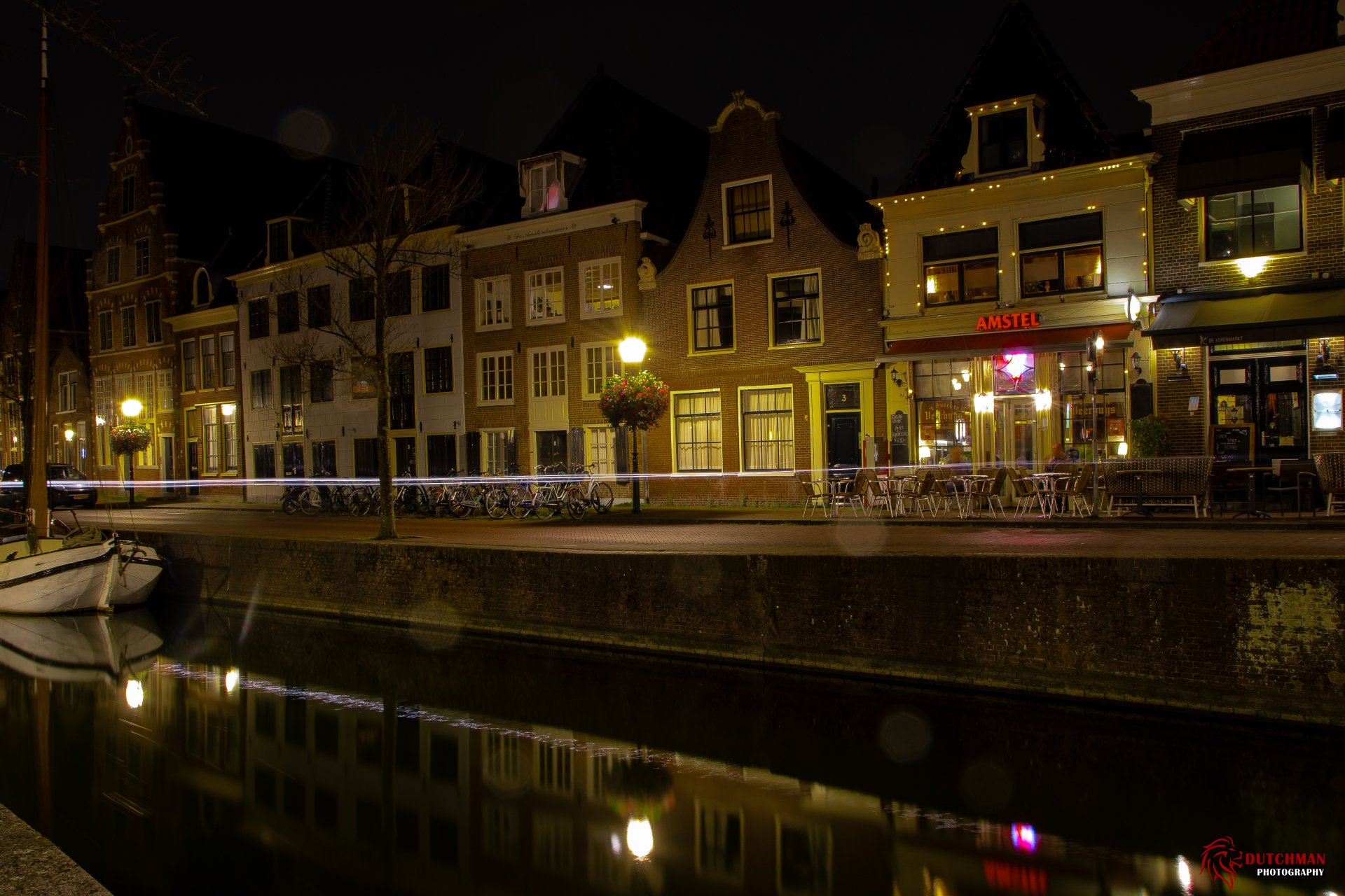 Night view of Hoorn’s waterfront with illuminated historic buildings, a lamp post, and their reflections on calm water in this 4K Ultra HD desktop wallpaper.