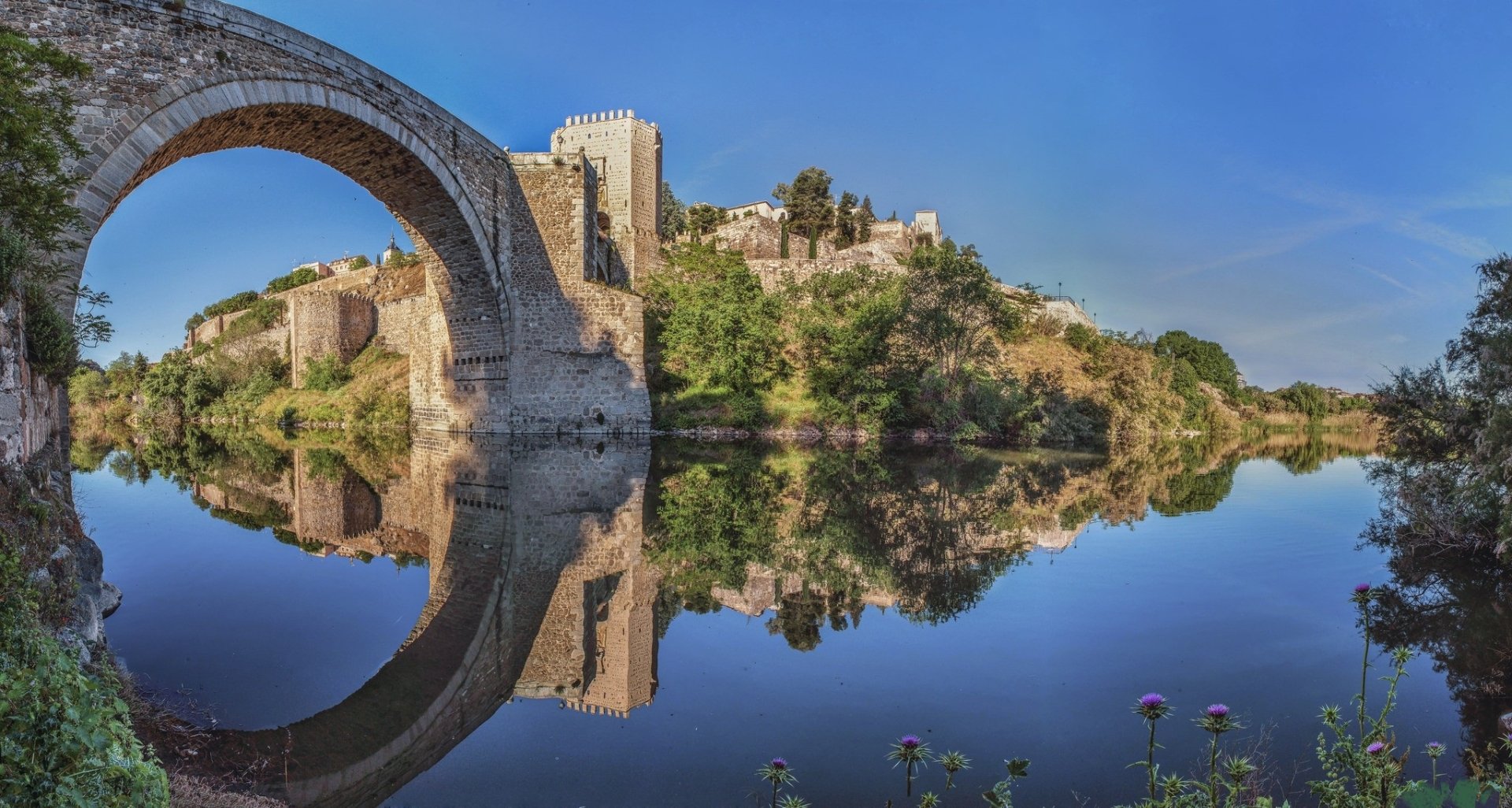 HD desktop wallpaper showcasing the Puente de Alcántara, a historic man-made stone bridge in Toledo, reflected beautifully in calm river waters under a clear blue sky.