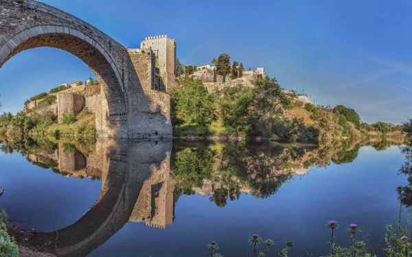 HD desktop wallpaper showcasing the Puente de Alcántara, a historic man-made stone bridge in Toledo, reflected beautifully in calm river waters under a clear blue sky.