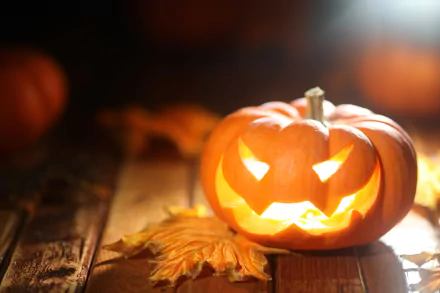 Glowing jack-o'-lantern on a wooden surface with autumn leaves, capturing a festive Halloween holiday vibe in stunning 4K Ultra HD for PC desktop wallpaper.