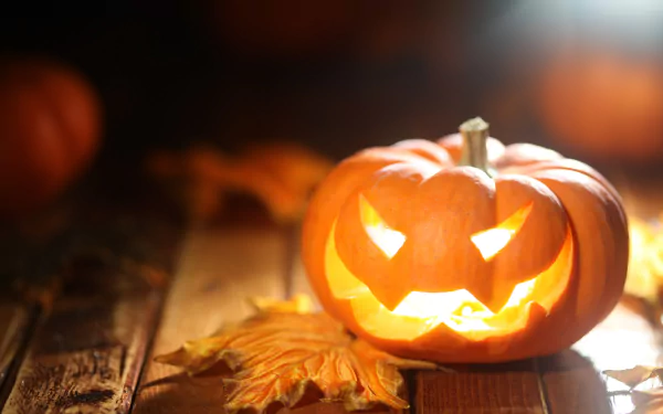 Glowing jack-o'-lantern on a wooden surface with autumn leaves, capturing a festive Halloween holiday vibe in stunning 4K Ultra HD for PC desktop wallpaper.