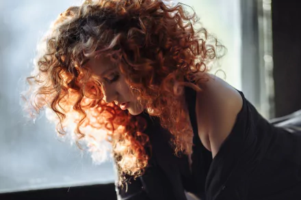2K Quad HD PC desktop wallpaper: moody redhead model with curly hair gazing downward by a window, wearing an off-shoulder black top.
