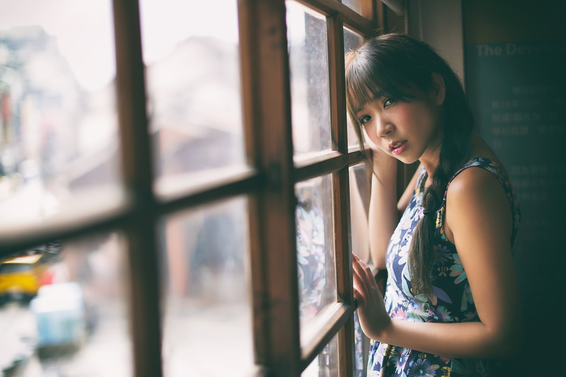 A HD desktop wallpaper featuring an Asian brunette woman with a braid leaning against a window, looking thoughtfully outside.