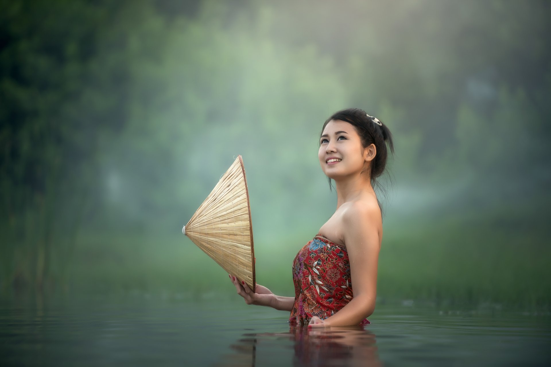 5K Ultra HD desktop wallpaper: smiling woman model with black hair waist-deep in water holding a conical hat, shallow depth of field and soft green background.