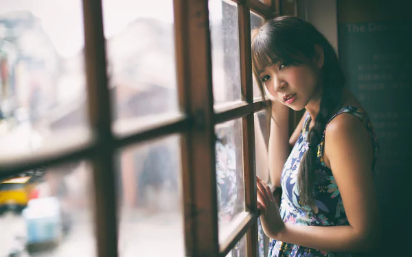 A HD desktop wallpaper featuring an Asian brunette woman with a braid leaning against a window, looking thoughtfully outside.