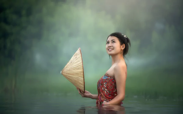 5K Ultra HD desktop wallpaper: smiling woman model with black hair waist-deep in water holding a conical hat, shallow depth of field and soft green background.
