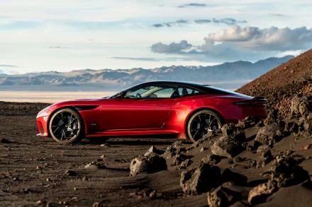 Red Aston Martin DBS Superleggera supercar parked on rocky terrain with mountains and clouds in the background, captured as an HD PC desktop wallpaper.