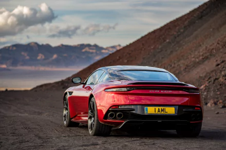 HD desktop wallpaper featuring a red Aston Martin DBS Superleggera supercar parked on a scenic mountainous road under a cloudy sky.