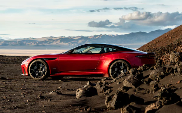 Red Aston Martin DBS Superleggera supercar parked on rocky terrain with mountains and clouds in the background, captured as an HD PC desktop wallpaper.