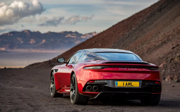 HD desktop wallpaper featuring a red Aston Martin DBS Superleggera supercar parked on a scenic mountainous road under a cloudy sky.