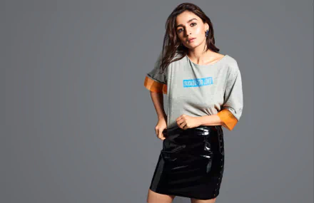 Brunette woman with brown eyes posed against a gray backdrop, wearing a gray top with orange cuffs and a black patent skirt — HD PC desktop wallpaper.