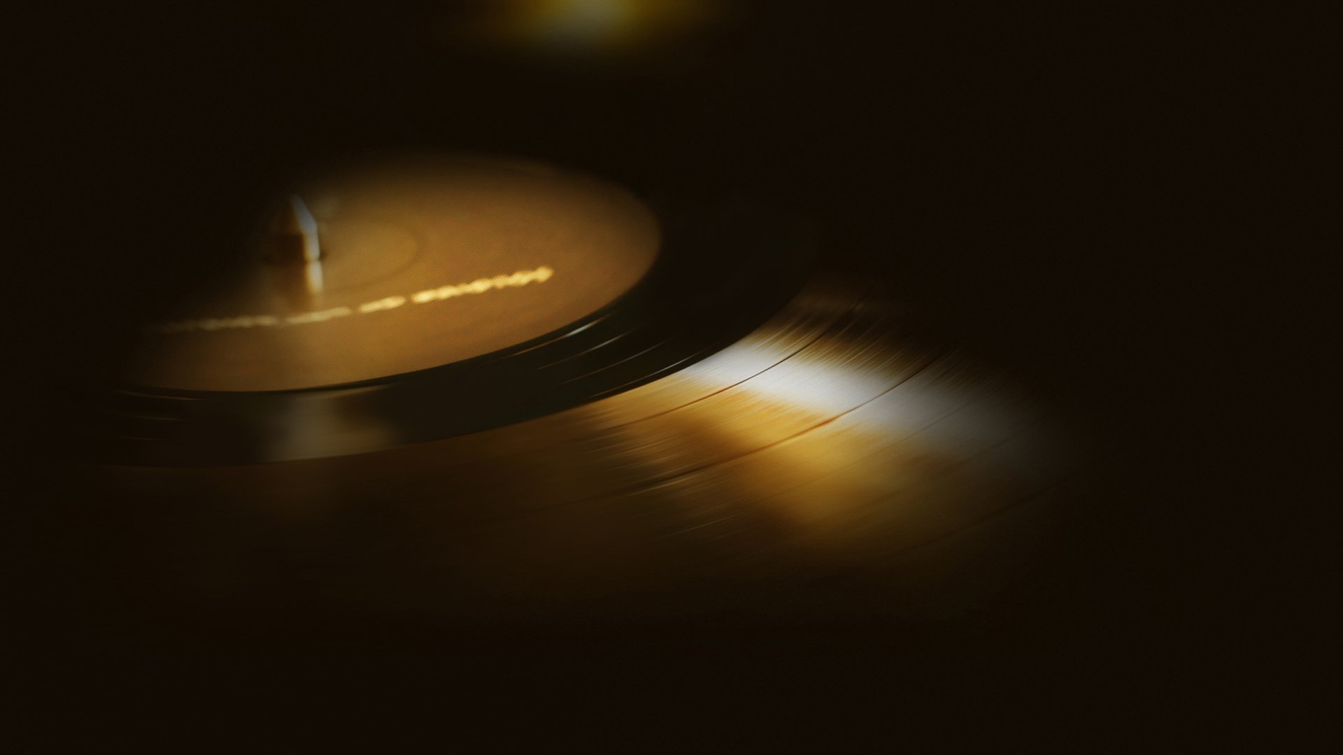 Close-up vinyl record on a turntable with warm golden reflections and deep shadows — moody 2K Quad HD PC desktop wallpaper background capturing analog music.