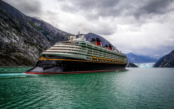 HD desktop wallpaper featuring the Disney Wonder cruise ship sailing through a serene fjord surrounded by rugged mountains under a cloudy sky.