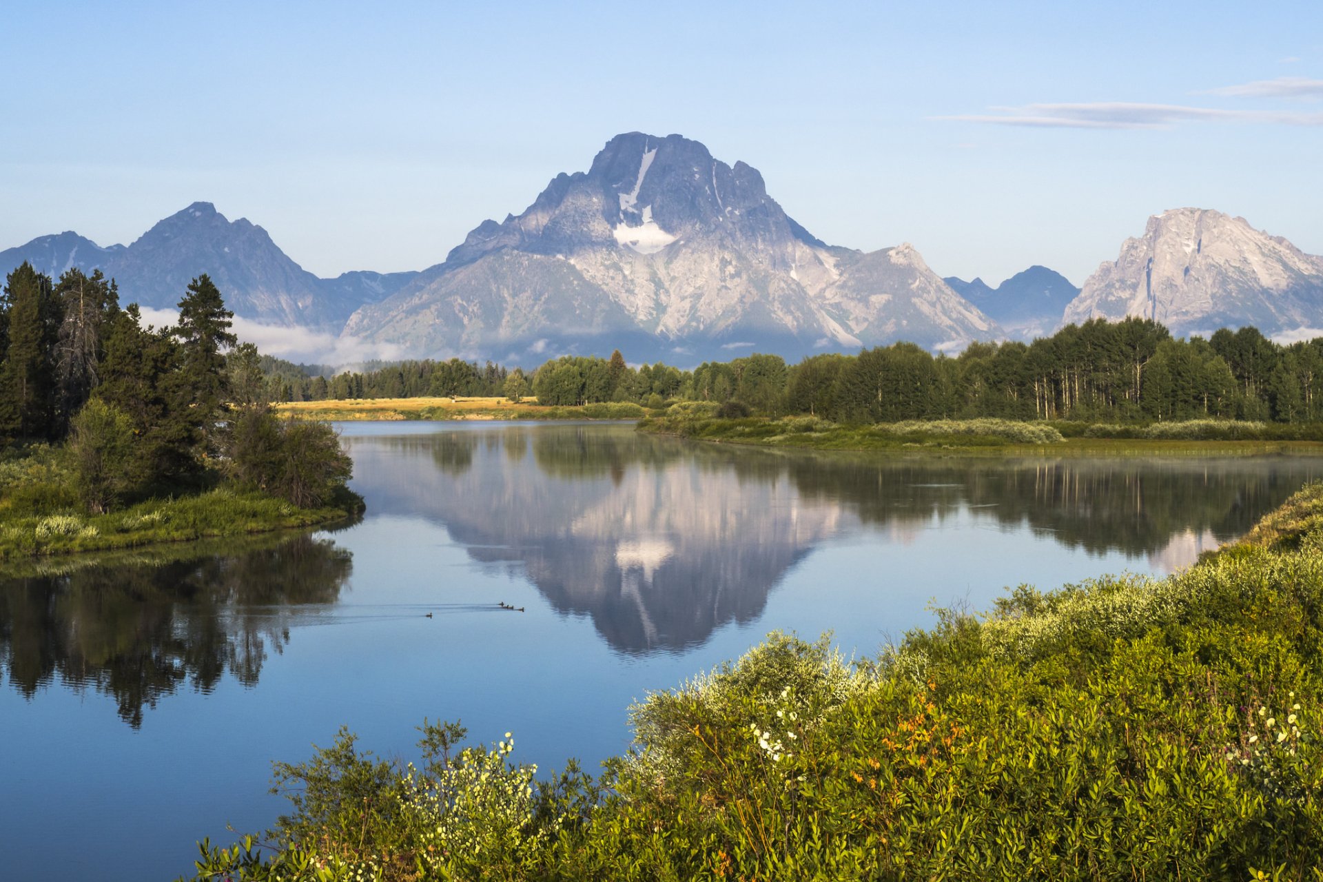HD desktop wallpaper of a serene lake reflecting the majestic peaks of Grand Teton National Park, surrounded by lush greenery and clear blue skies.