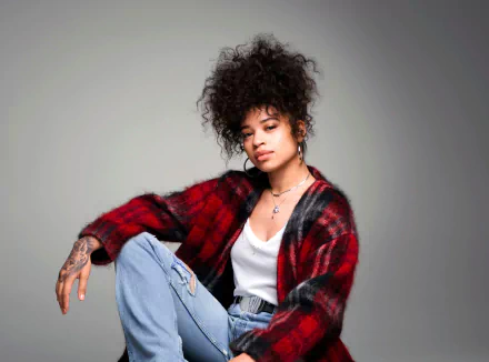 HD desktop wallpaper of Ella Mai seated in a casual pose, wearing a red flannel and jeans against a gray background.