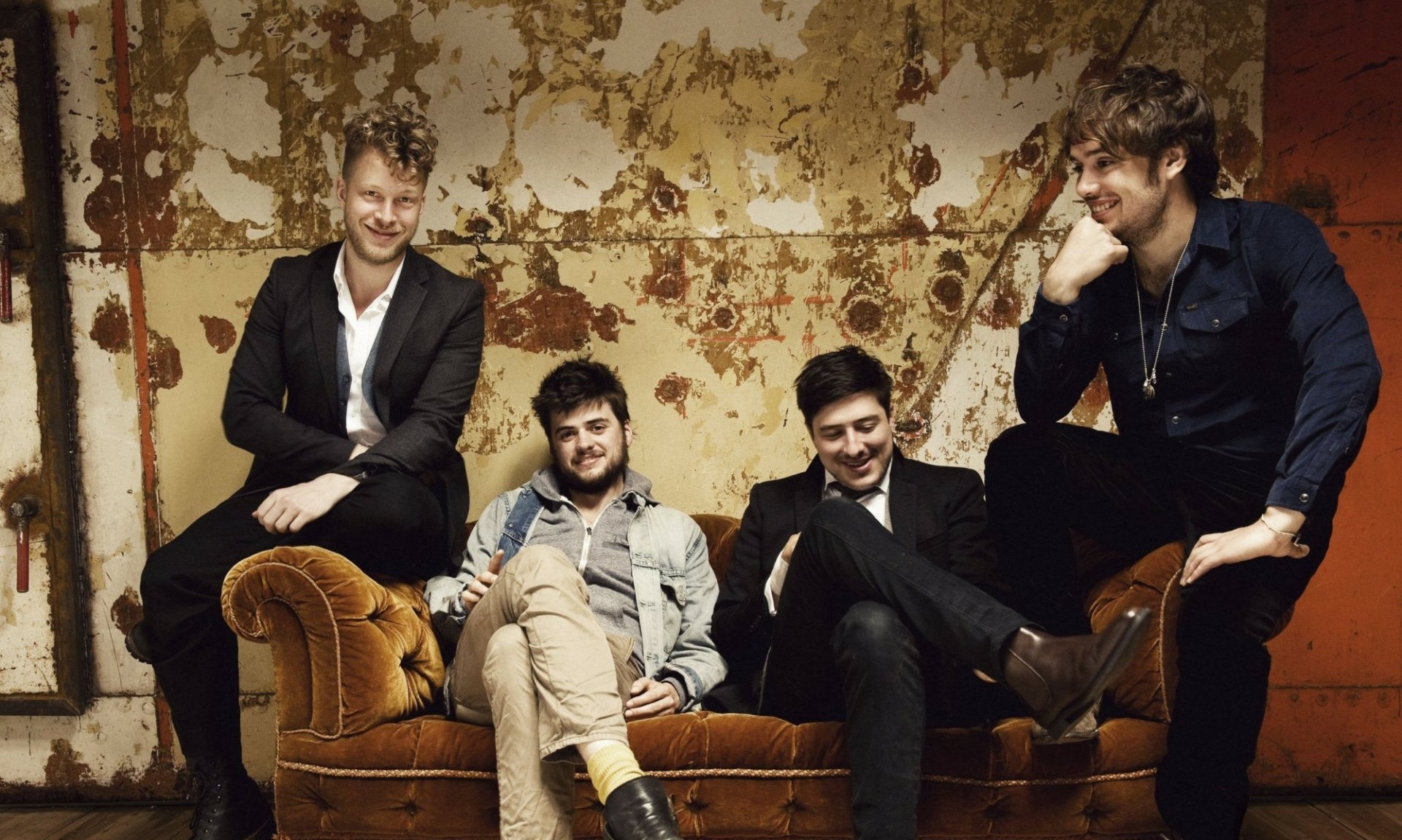 HD wallpaper of Mumford & Sons band members casually seated and leaning against a wall with peeling paint, exuding a relaxed and vintage vibe.
