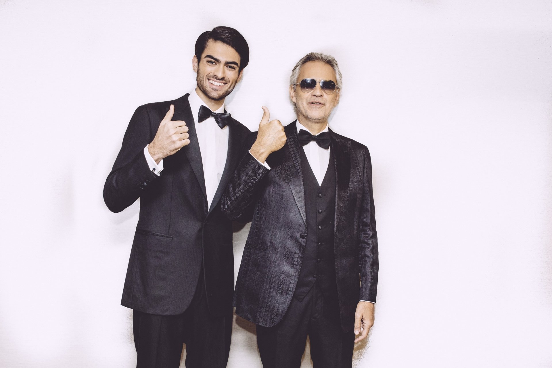 HD desktop wallpaper featuring Andrea Bocelli and another artist, both dressed in stylish tuxedos, smiling and giving a thumbs-up against a simple background.