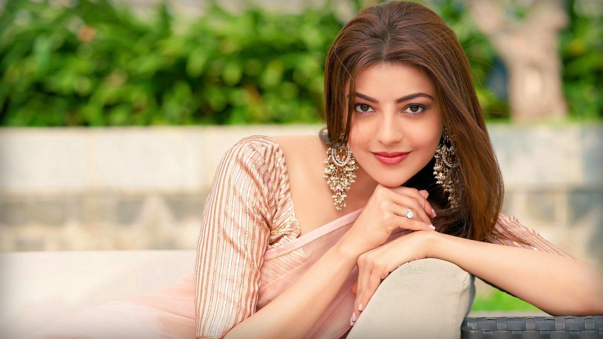 HD PC desktop wallpaper featuring celebrity Kajal Aggarwal in a soft pink outfit, adorned with statement earrings, posing outdoors with a green blurred background.