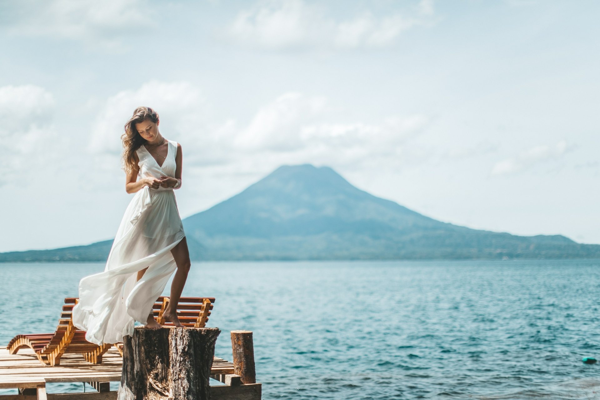 A blonde woman with long hair in a flowing white dress stands on a dock by a lake, with a mountain and cloudy sky in the background, captured in 4K Ultra HD.