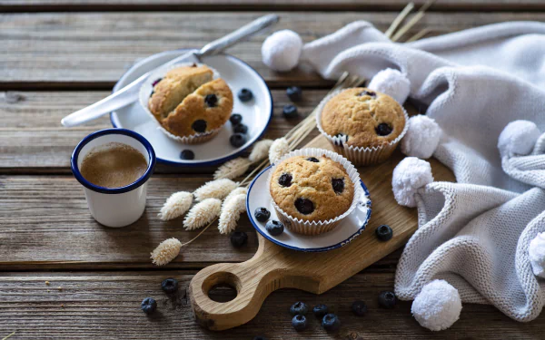 A still life 4K Ultra HD image of blueberry muffins on plates, accompanied by a cup of coffee and cozy white textiles on a wooden surface.