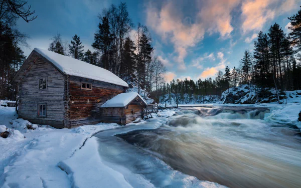 HD desktop wallpaper of a snowy cabin in a forest by a flowing river during winter in Finland, showcasing serene nature and tranquil winter scenery at dusk.