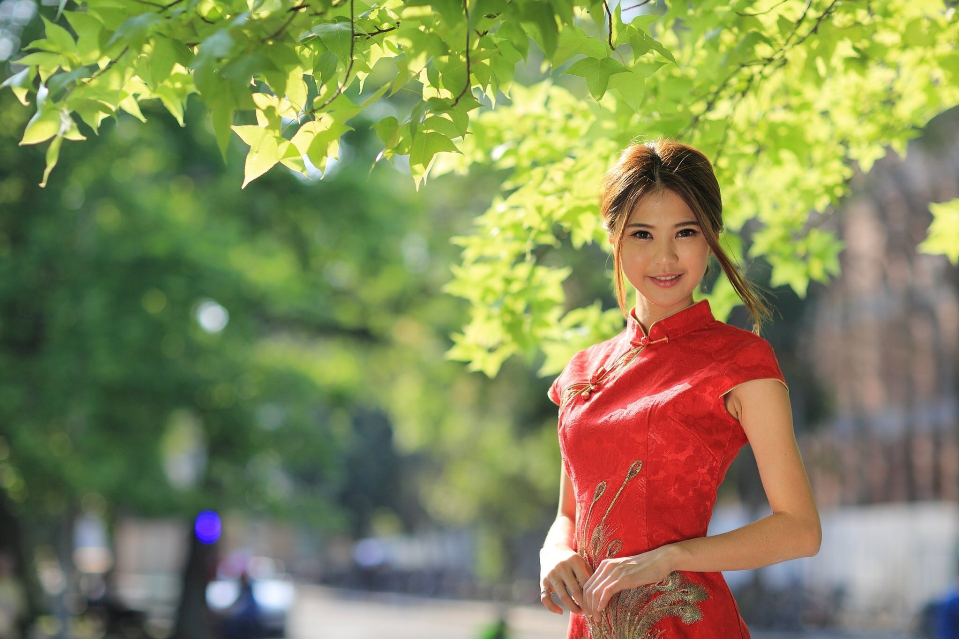 A smiling brunette woman with brown eyes, wearing a kimono, stands outdoors with blurred greenery in the background. The image has a depth of field effect and is an HD desktop wallpaper or background.