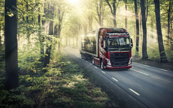 HD wallpaper featuring a Volvo truck driving on a forest road, with sunbeams filtering through the tall trees.